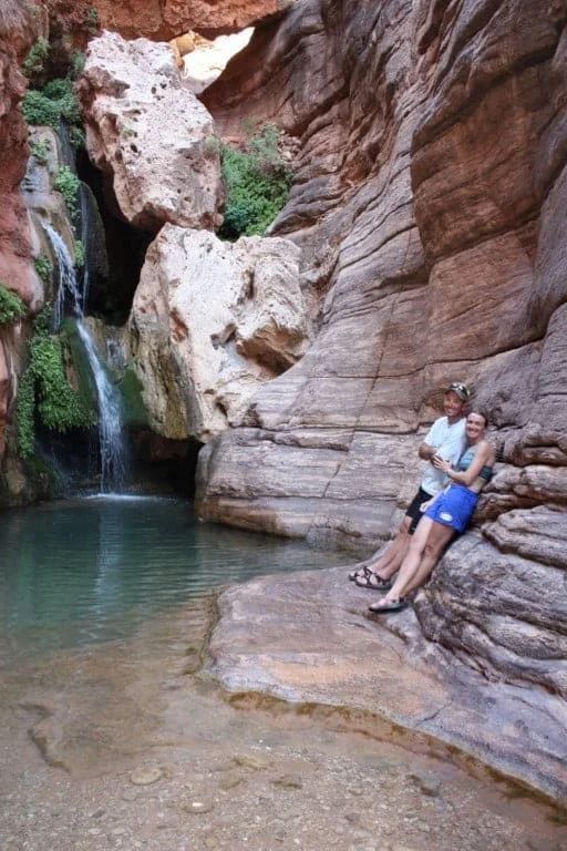 Waterfall cascading into a turquoise pool, with two people sitting near the water's edge in a red-rock canyon.