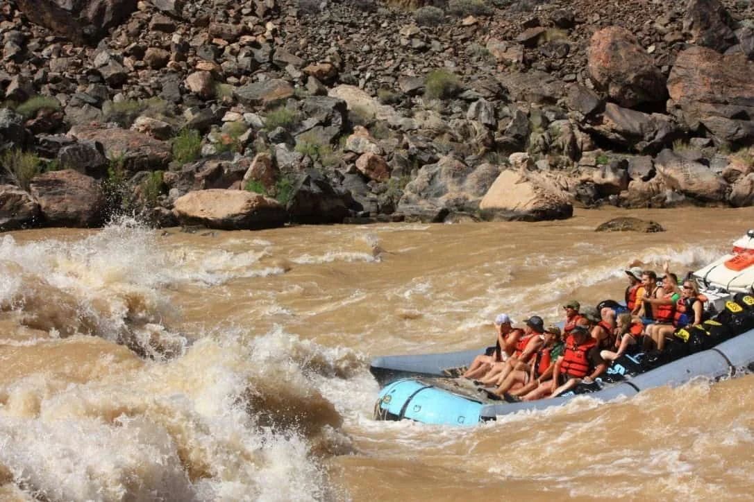 Raft with passengers navigates churning, muddy river near rocky canyon walls; passengers wear life vests.