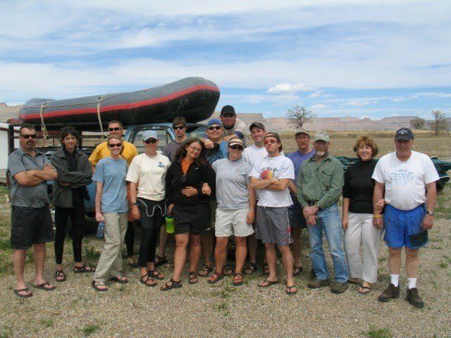 Group of people posing by a truck with a raft on top; outdoors with cloudy sky.