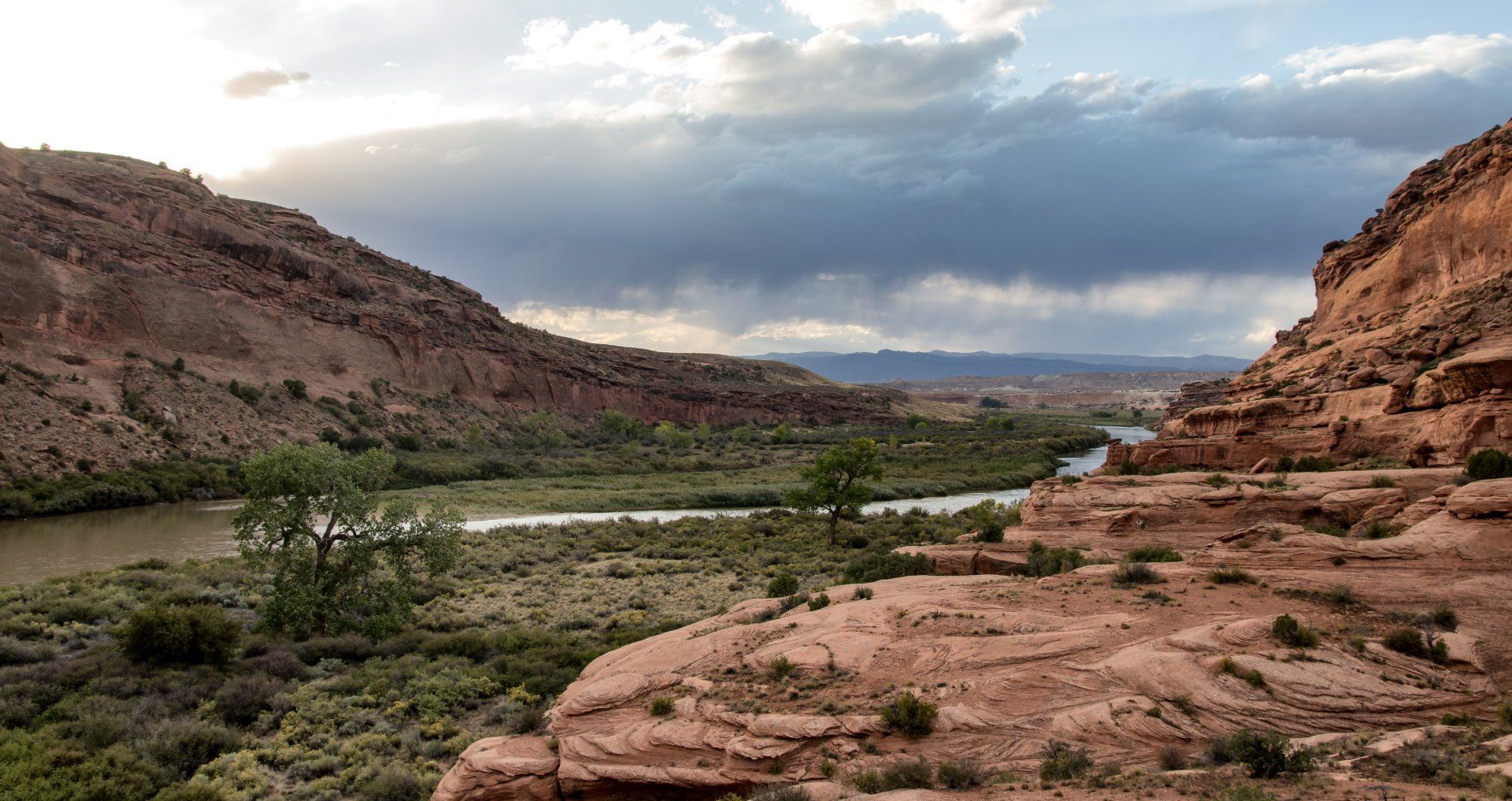 Canyon landscape with river, red rock cliffs, and cloudy sky.