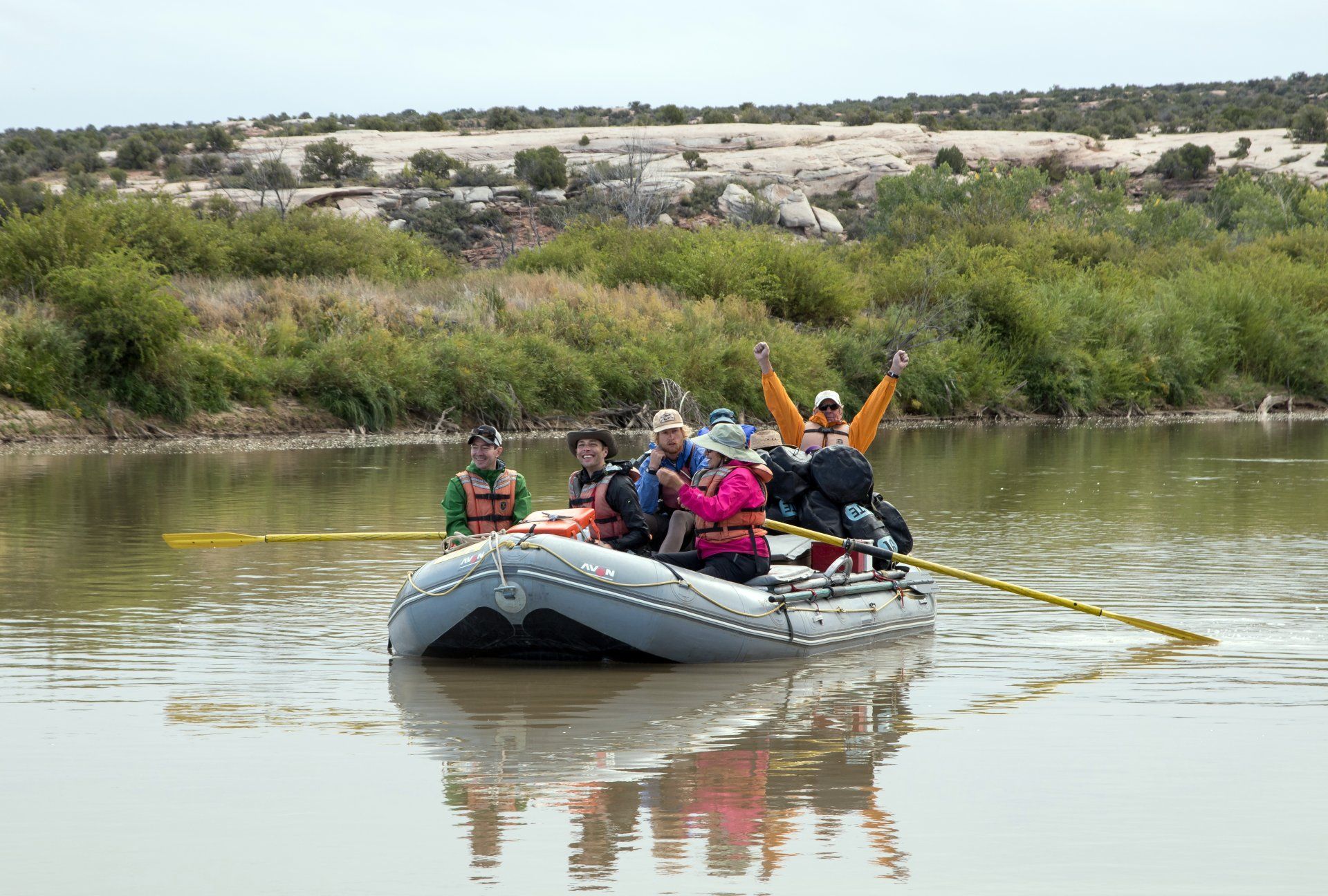 People in a raft on a river. One person raises arms. Green foliage, desert landscape in background.