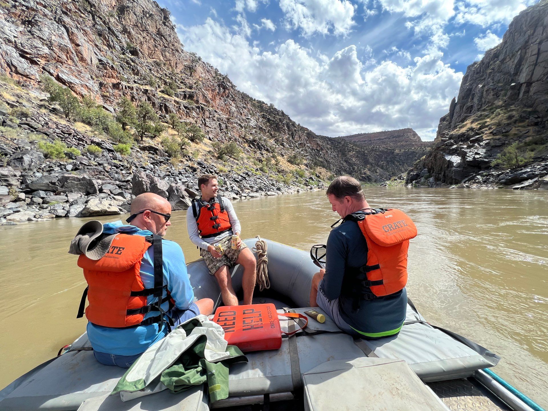 Three people in orange life vests on a raft navigate a river through a canyon. Cloudy sky.