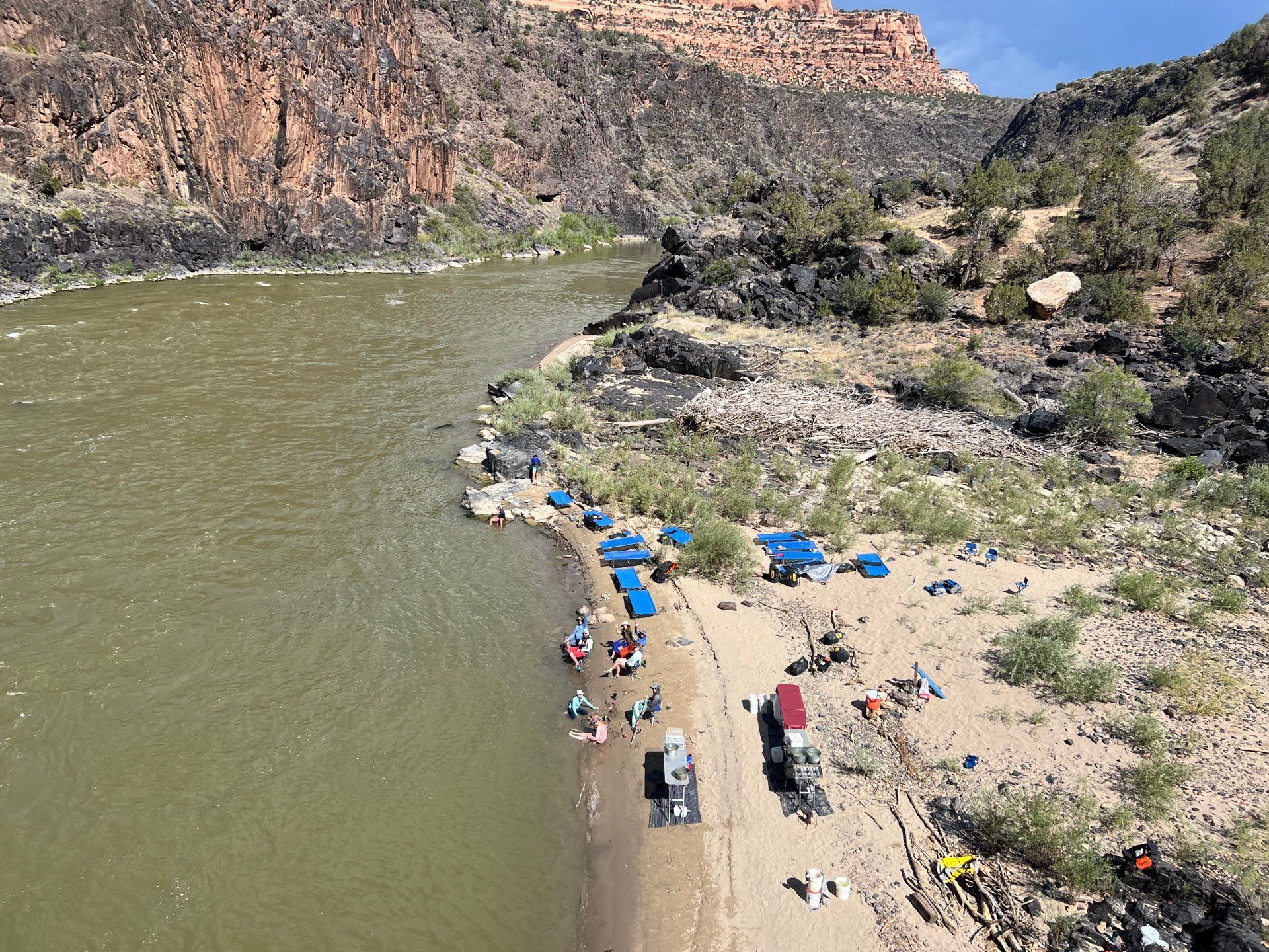 Riverbank campsite with tents, boats, and people near a cliffside.