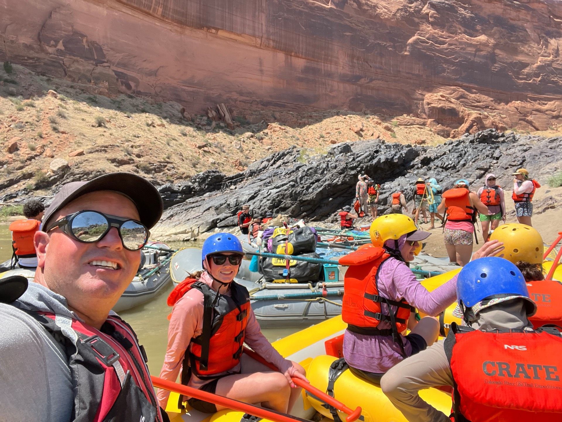 Group on rafts near a canyon wall, preparing for rafting. People wearing life vests and helmets.
