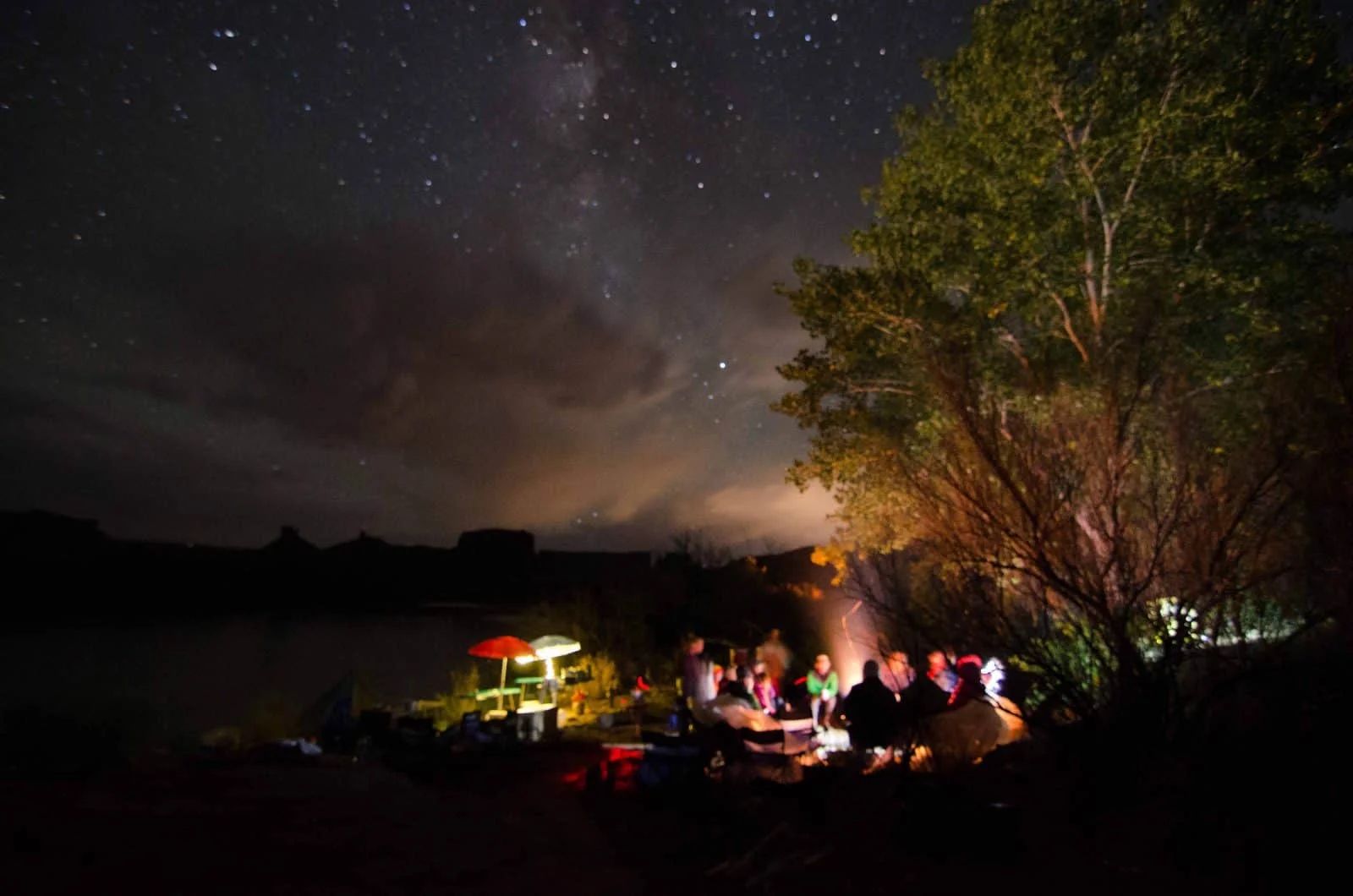Campfire gathering under a starry night sky near a body of water; silhouettes around firelight.