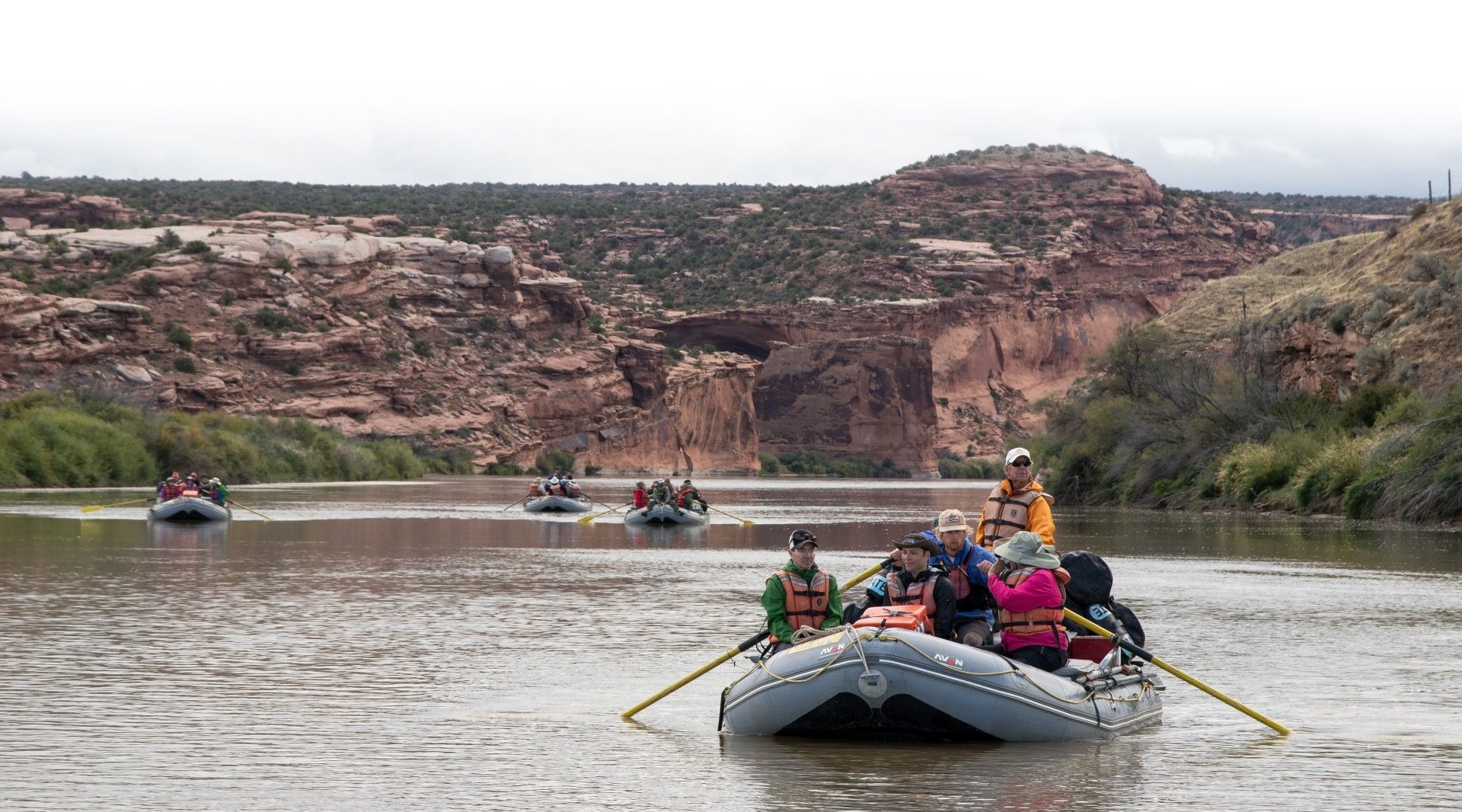 Rafts with people rowing on a river near rocky cliffs on an overcast day.