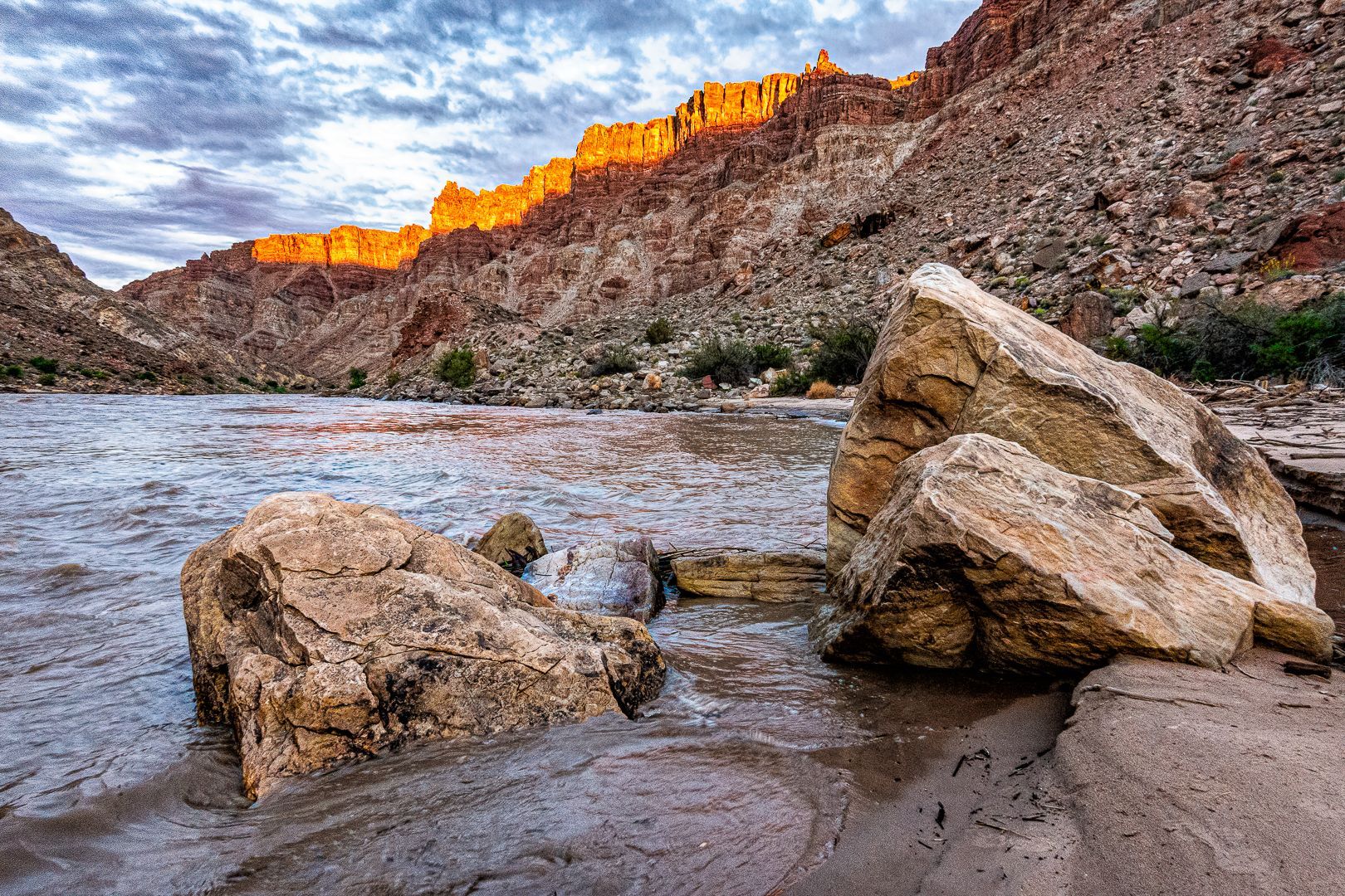 River flowing past large rocks in a canyon, with sunlit cliffs and cloudy sky.