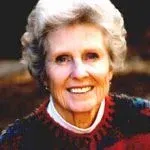 Woman with short white hair, smiling, wearing a red and blue sweater.