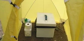 Inside a yellow portable toilet tent, a white portable toilet with a toilet paper holder.