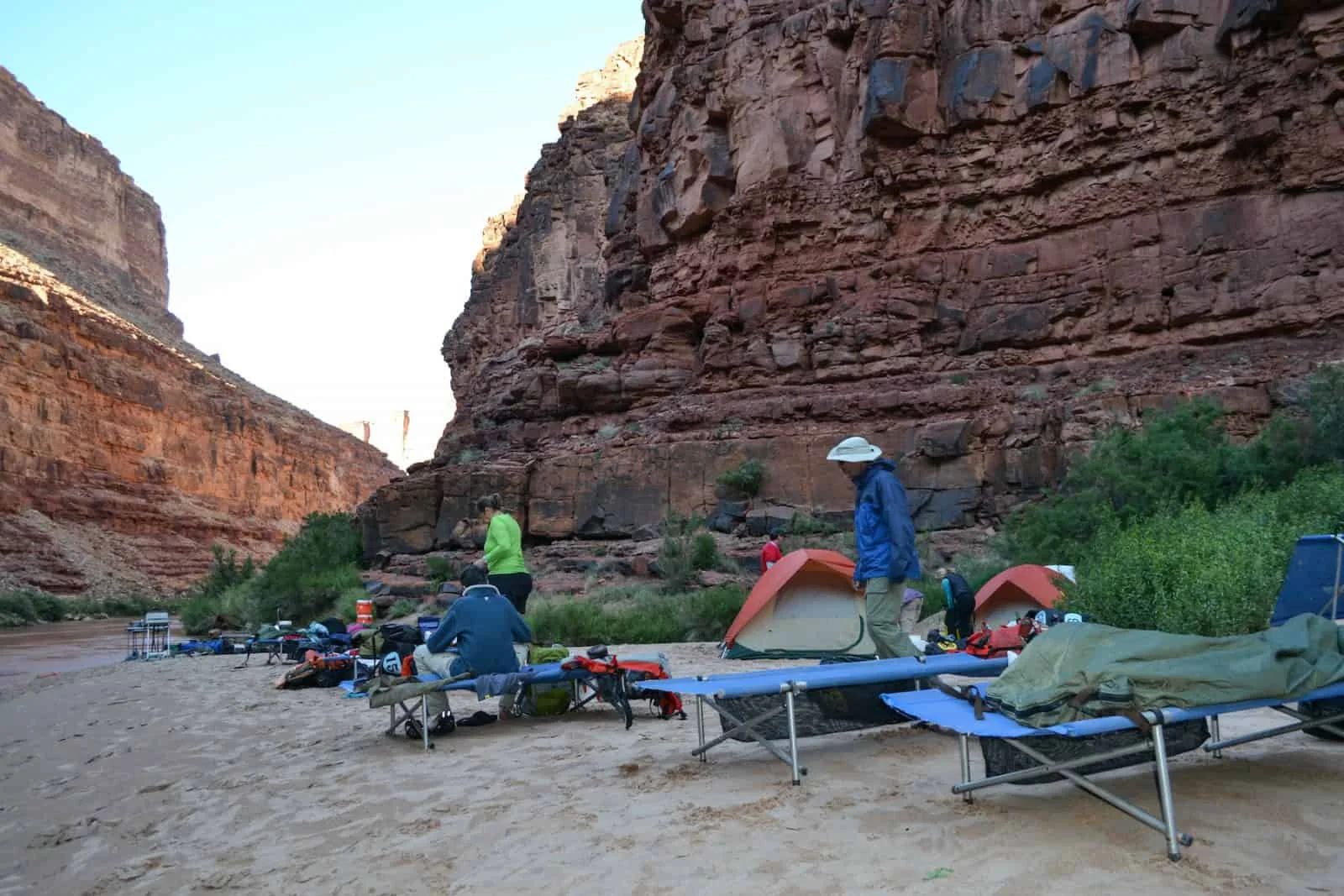 Campers with tents and cots set up on a sandy beach at the base of red rock canyon walls.