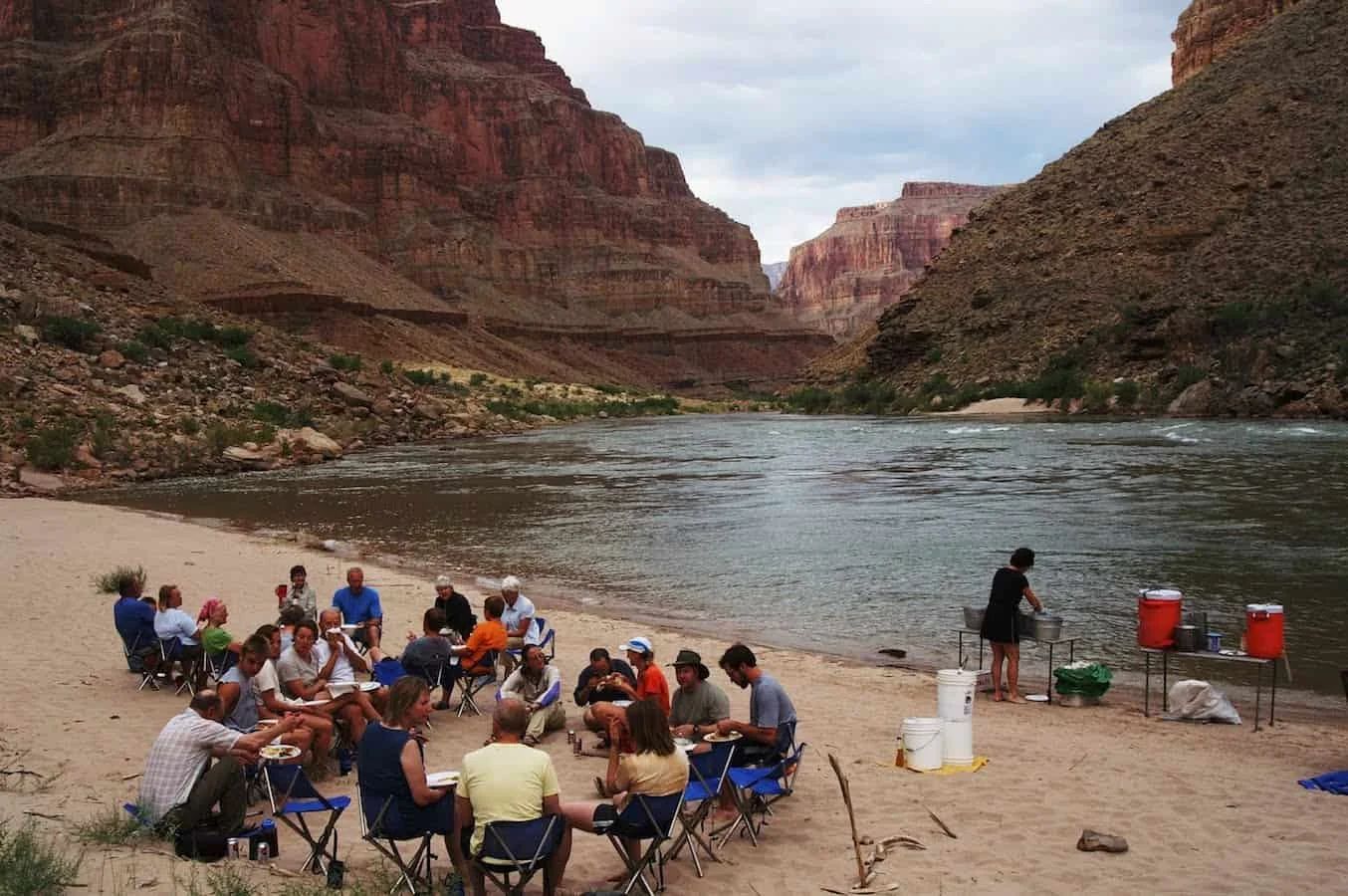 People gathered on a sandy riverbank in a canyon, seated in folding chairs, eating with red-rock cliffs in the background.