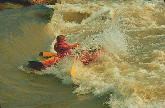 Kayaker in a red suit paddles through churning white water rapids, splashing.