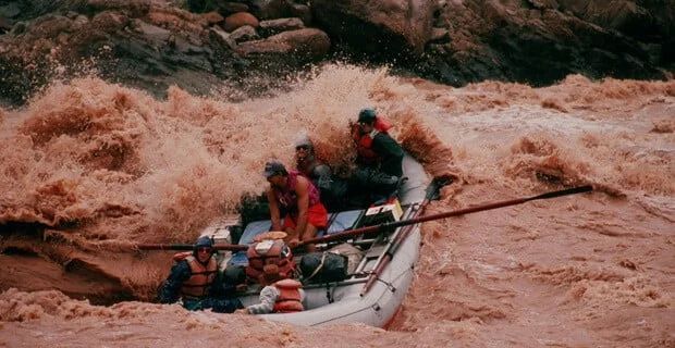 Raft navigating a raging, muddy river. People in life vests battle waves. Brown water splashes.