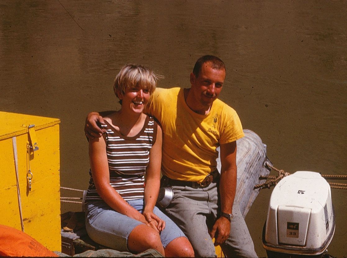 Couple in a boat, smiling. Woman in striped shirt, man in yellow shirt, arm around her. Yellow box. Brown water background.