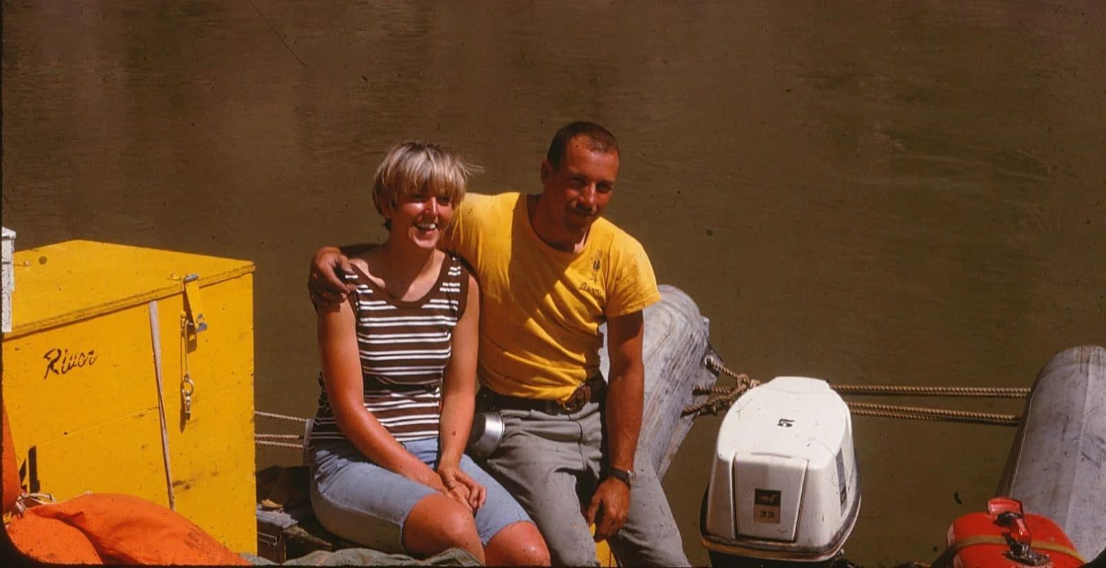 Couple on boat smiling, arm around each other. Water and yellow box in background.