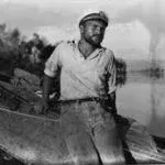 Man in cap and short sleeves leans on wood, near water.