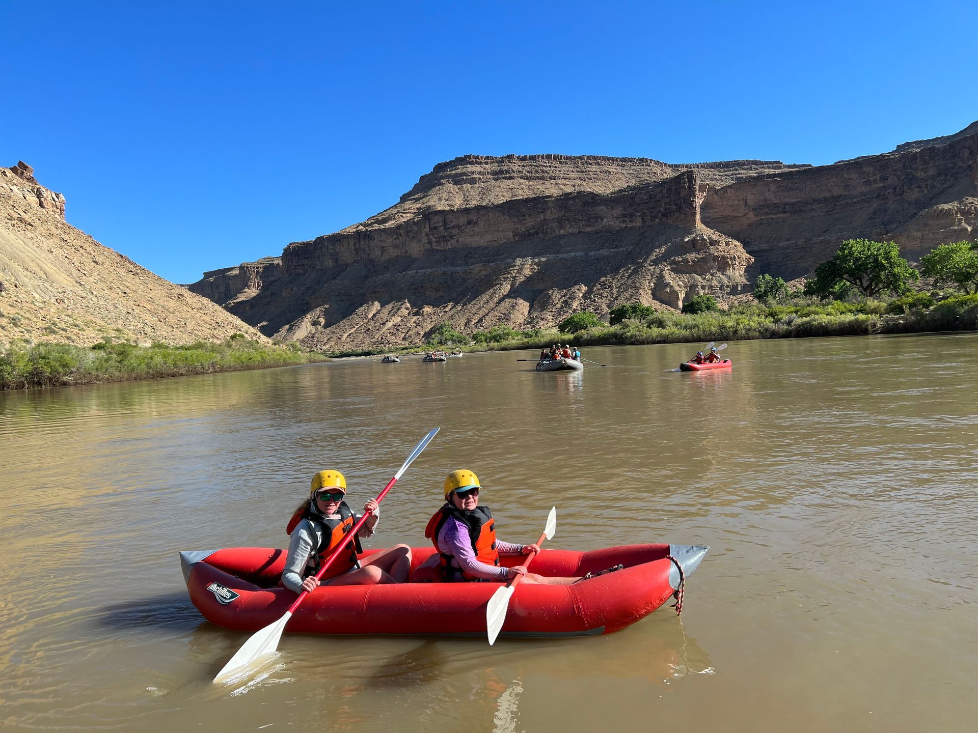 Two people in a red raft paddle on a brown river, with canyon walls in the background under a blue sky.