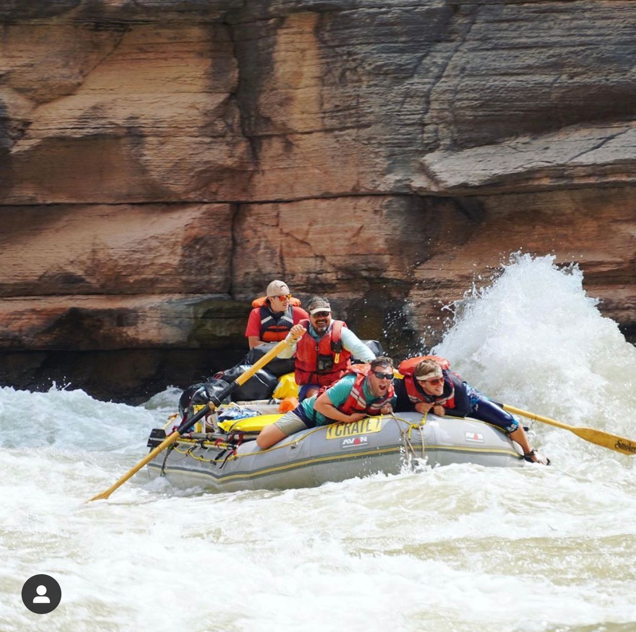 Rafting on a whitewater river; people brace against a wave. Brown rock wall in background.