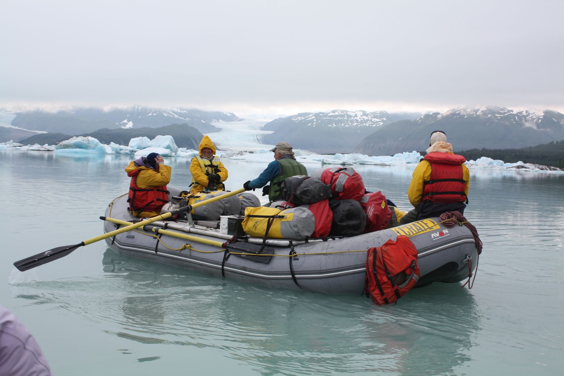 People in a raft on glacial lake, surrounded by icebergs. Overcast sky.