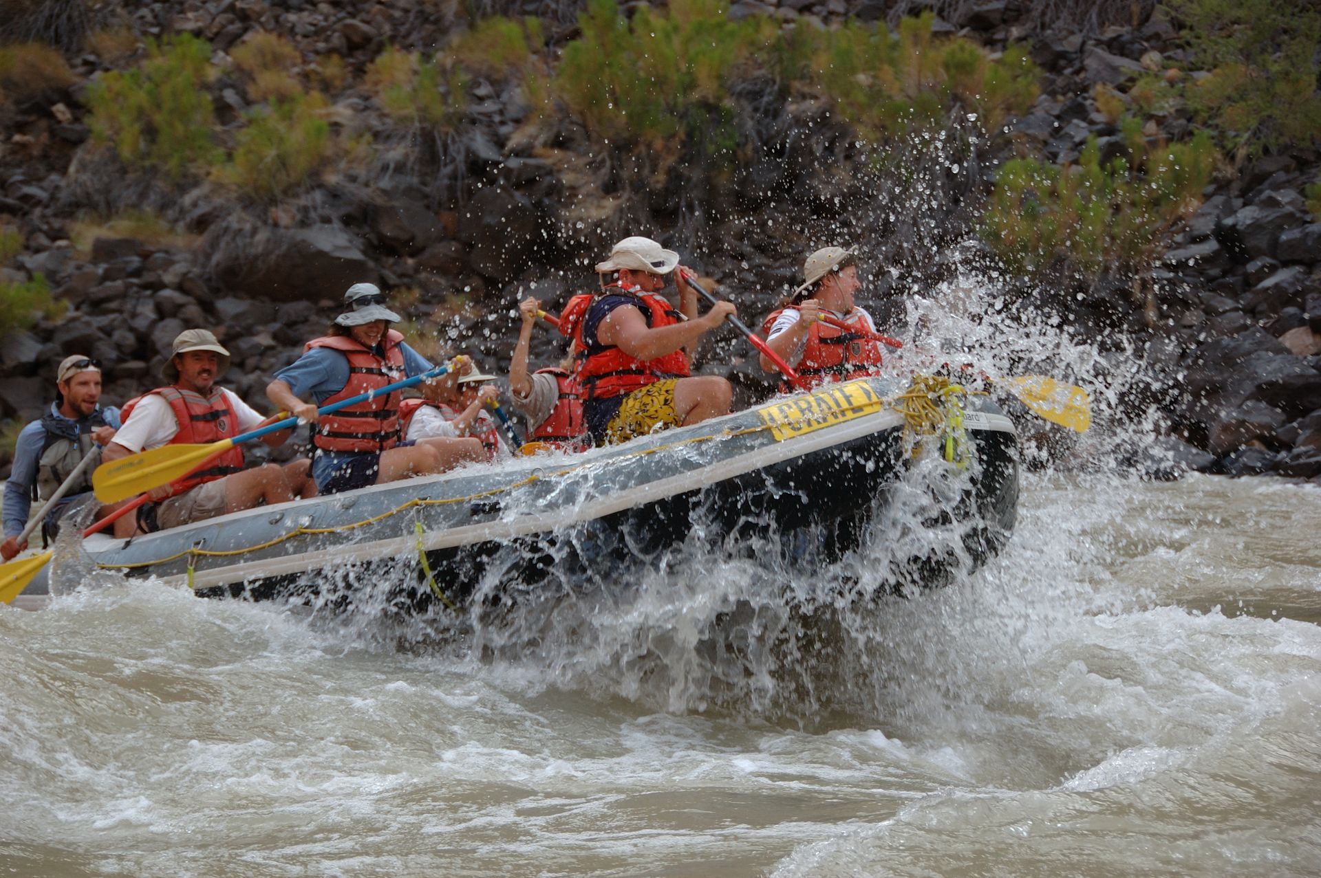 Whitewater rafters navigating a rapid, water splashing; river, rocks and foliage in background.