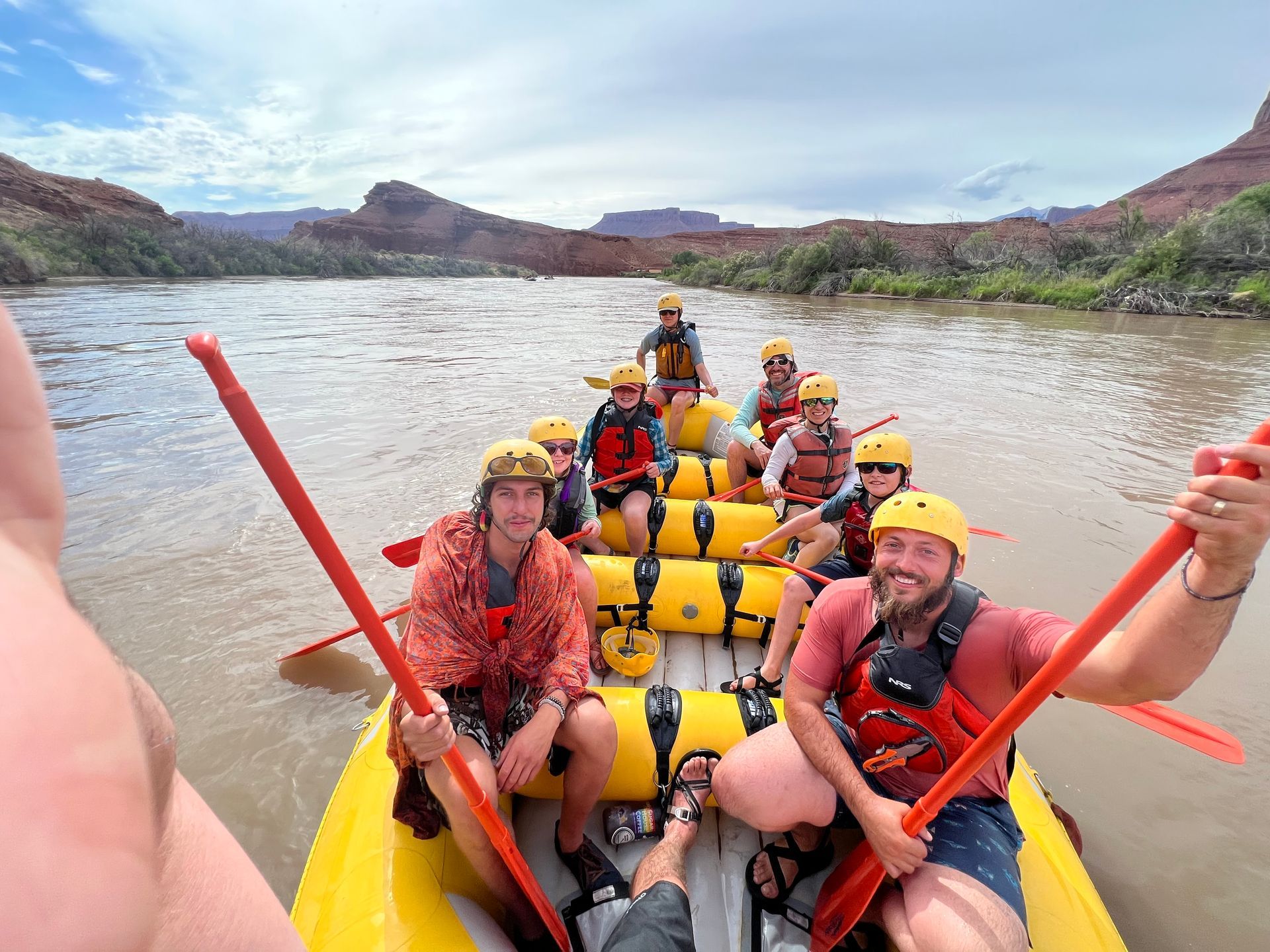 People rafting on a river, wearing helmets and life vests. Red paddles, yellow raft. Mountains in the background.
