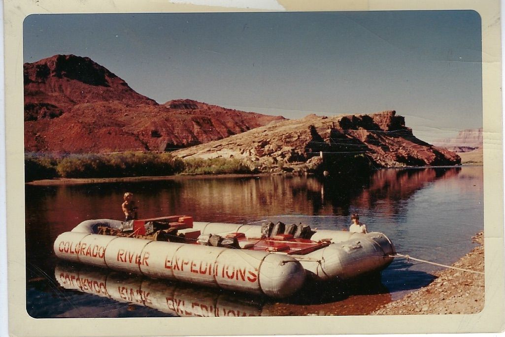 Rafting on calm river. Raft on shore, with red and brown desert landscape in background.