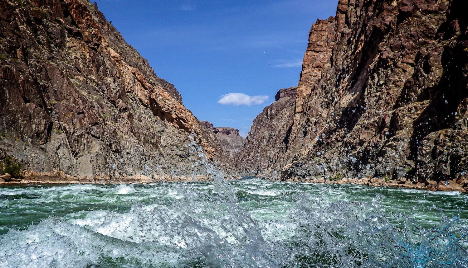 Raging river through a canyon with rocky cliffs and a blue sky. Water splashes in the foreground.