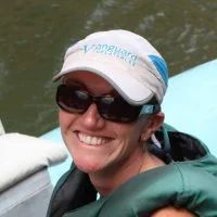 Woman wearing sunglasses and a cap smiles outdoors, likely on a boat.