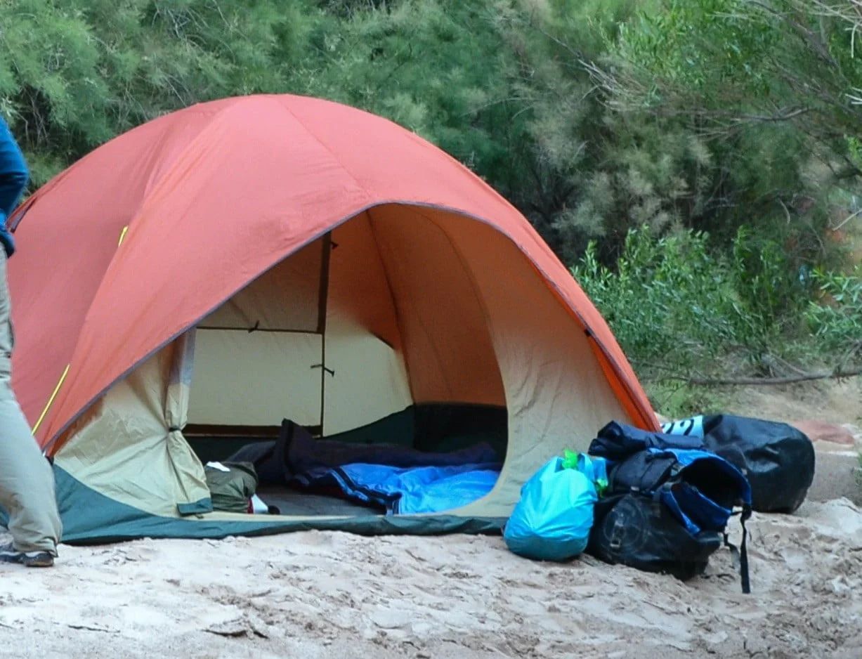 Red and tan camping tent set up outdoors, with sleeping bag inside. Backpacks sit outside.