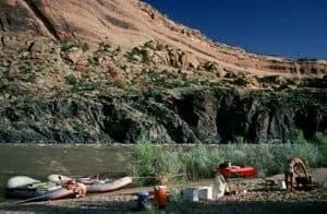 Rafts on riverbank with people preparing gear, red rock cliffs in background under blue sky.