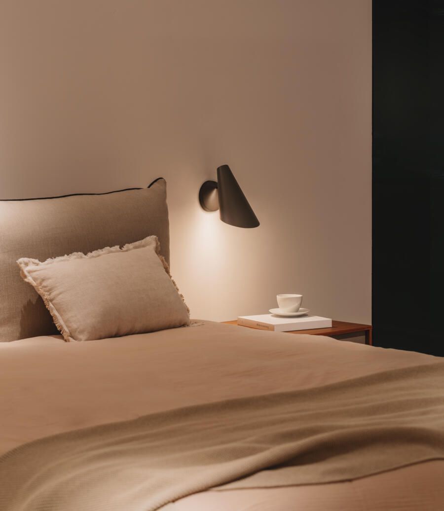 Un rincón de dormitorio minimalista con una cama beige con almohada, una pequeña mesita de noche de madera con una taza y una lámpara de pared.