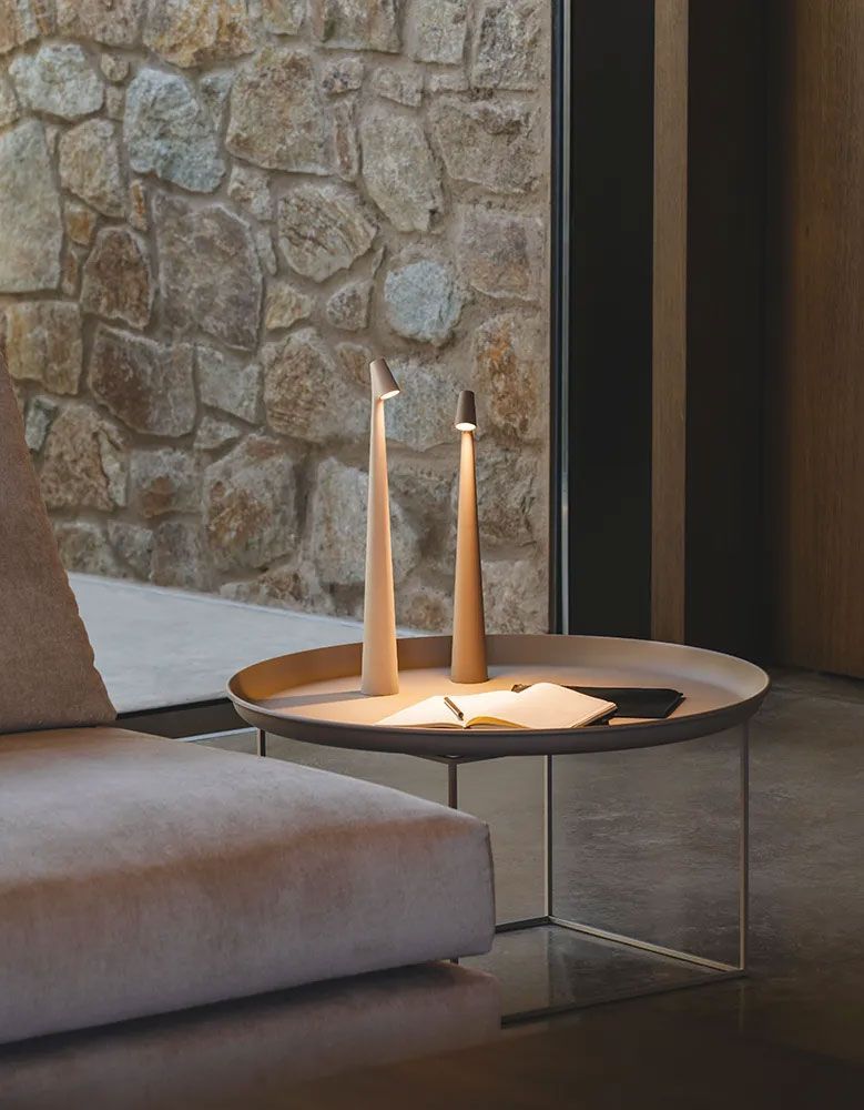 Dos lámparas de mesa delgadas y con forma de cono se encuentran sobre una mesa auxiliar circular junto a un sofá de color claro, frente a una pared de piedra.