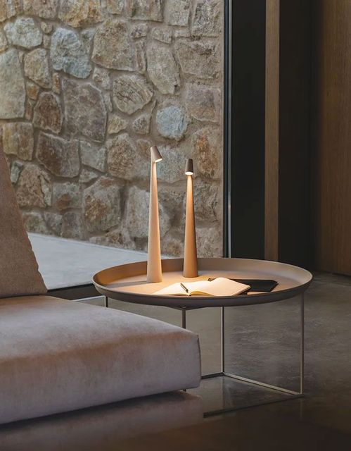 Dos lámparas de mesa delgadas y con forma de cono se encuentran sobre una mesa auxiliar circular junto a un sofá de color claro, frente a una pared de piedra.