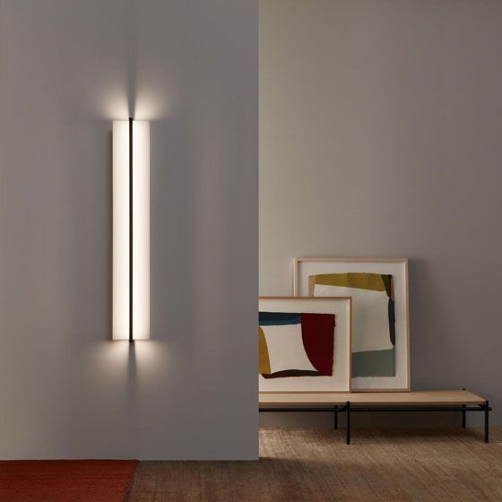 Un aplique de pared vertical iluminado ilumina un rincón donde se encuentra un cuadro de arte abstracto enmarcado sobre un banco bajo cercano.