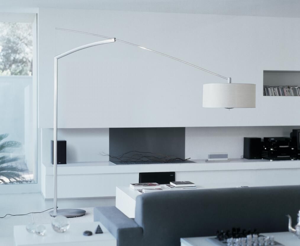 Una lámpara de pie minimalista de color blanco, con brazo arqueado y pantalla cilíndrica, preside un salón moderno decorado en tonos blancos.
