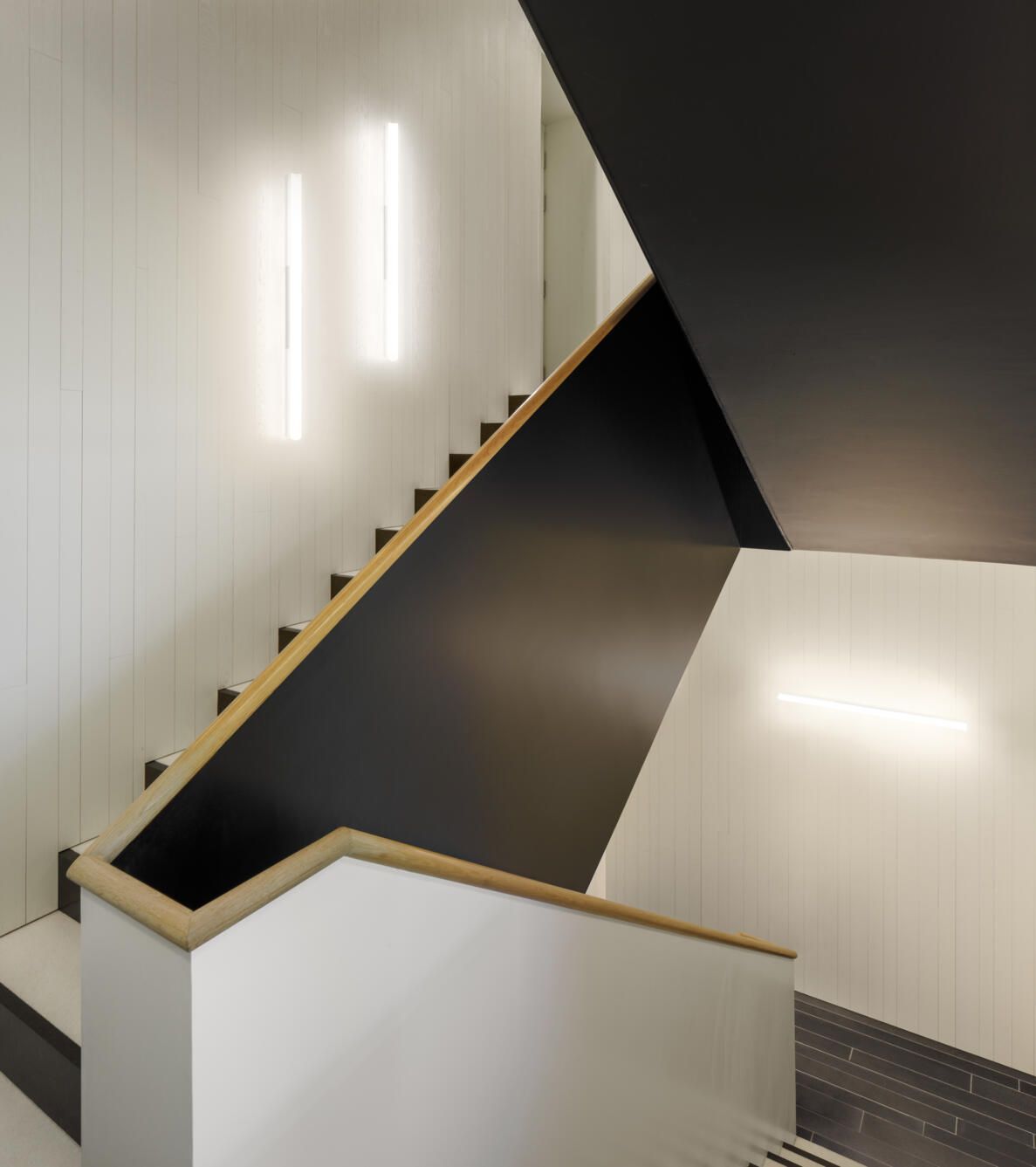 Una escalera moderna con paredes blancas, pasamanos negros y apliques de luz verticales en la pared.
