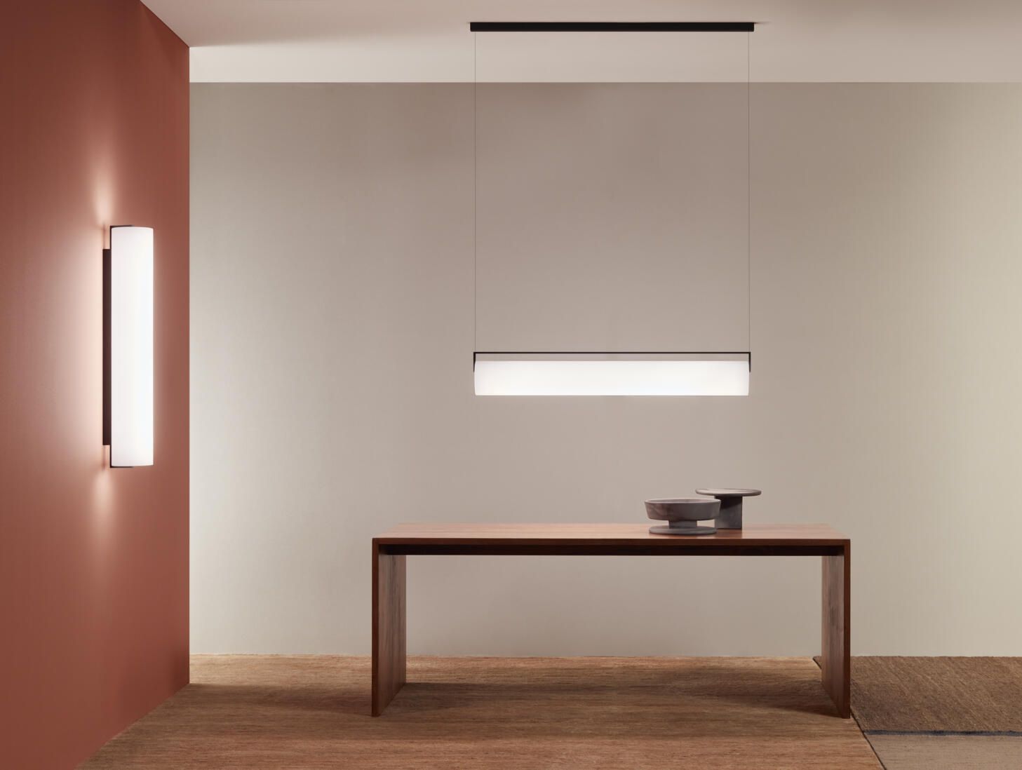 Una habitación moderna con una mesa de madera, una lámpara colgante lineal y un aplique de pared vertical a juego sobre una pared de terracota.