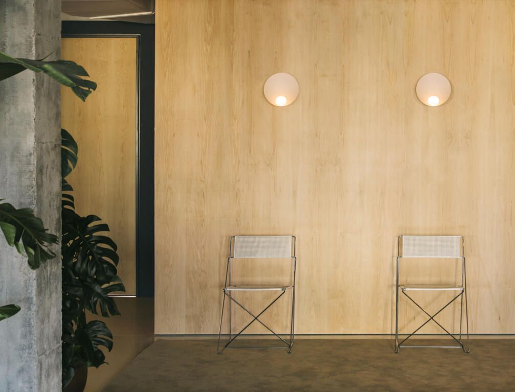 Dos sillas plegables minimalistas colocadas contra una pared revestida de madera clara, con dos apliques de pared circulares, en una habitación tranquila.