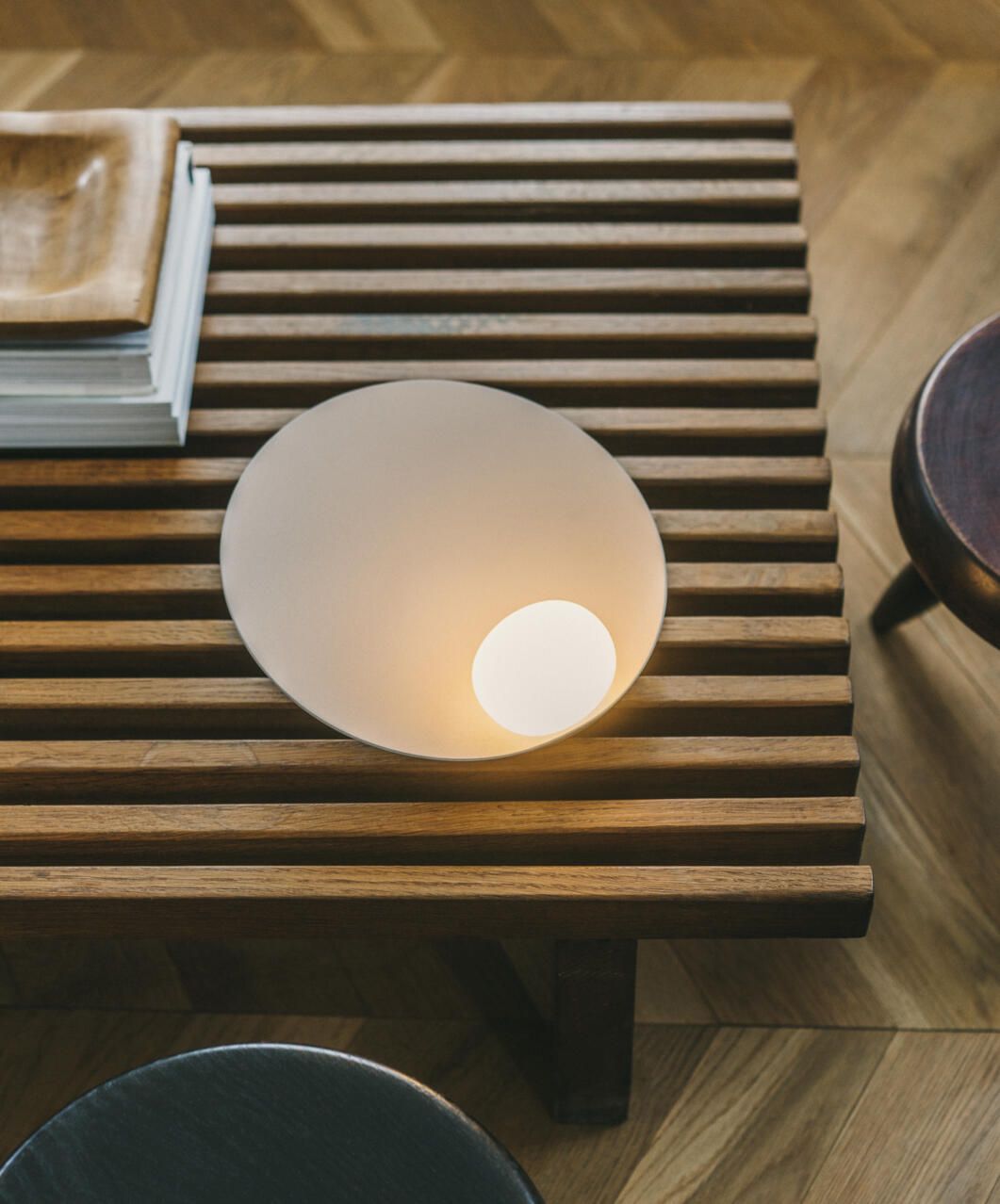 Una lámpara blanca circular y moderna ilumina un banco de madera con listones, con libros y taburetes cerca, sobre un suelo de madera.