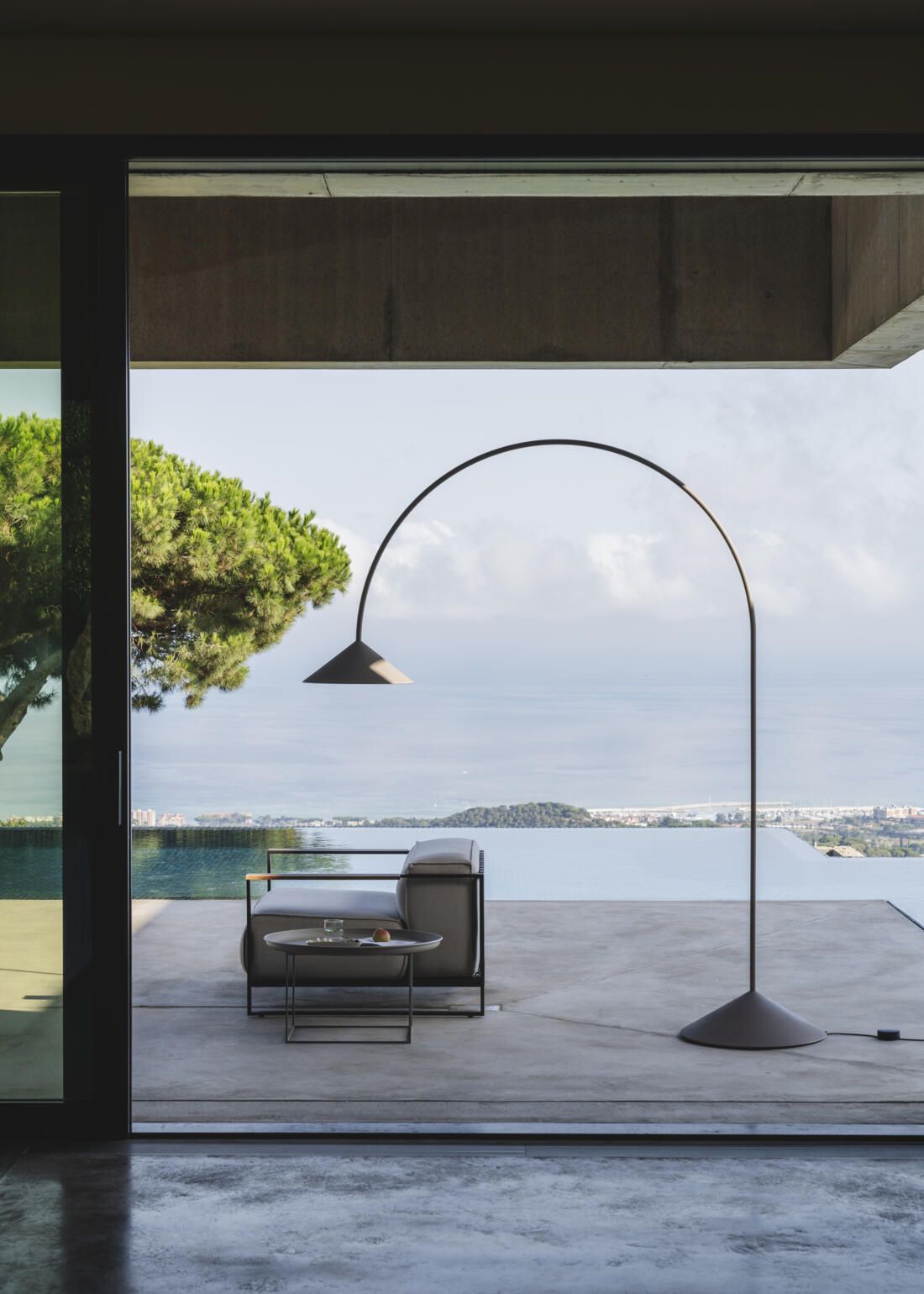 Una moderna lámpara de pie arqueada se alza sobre una terraza con vistas a un mar en calma y a la lejanía de la costa.