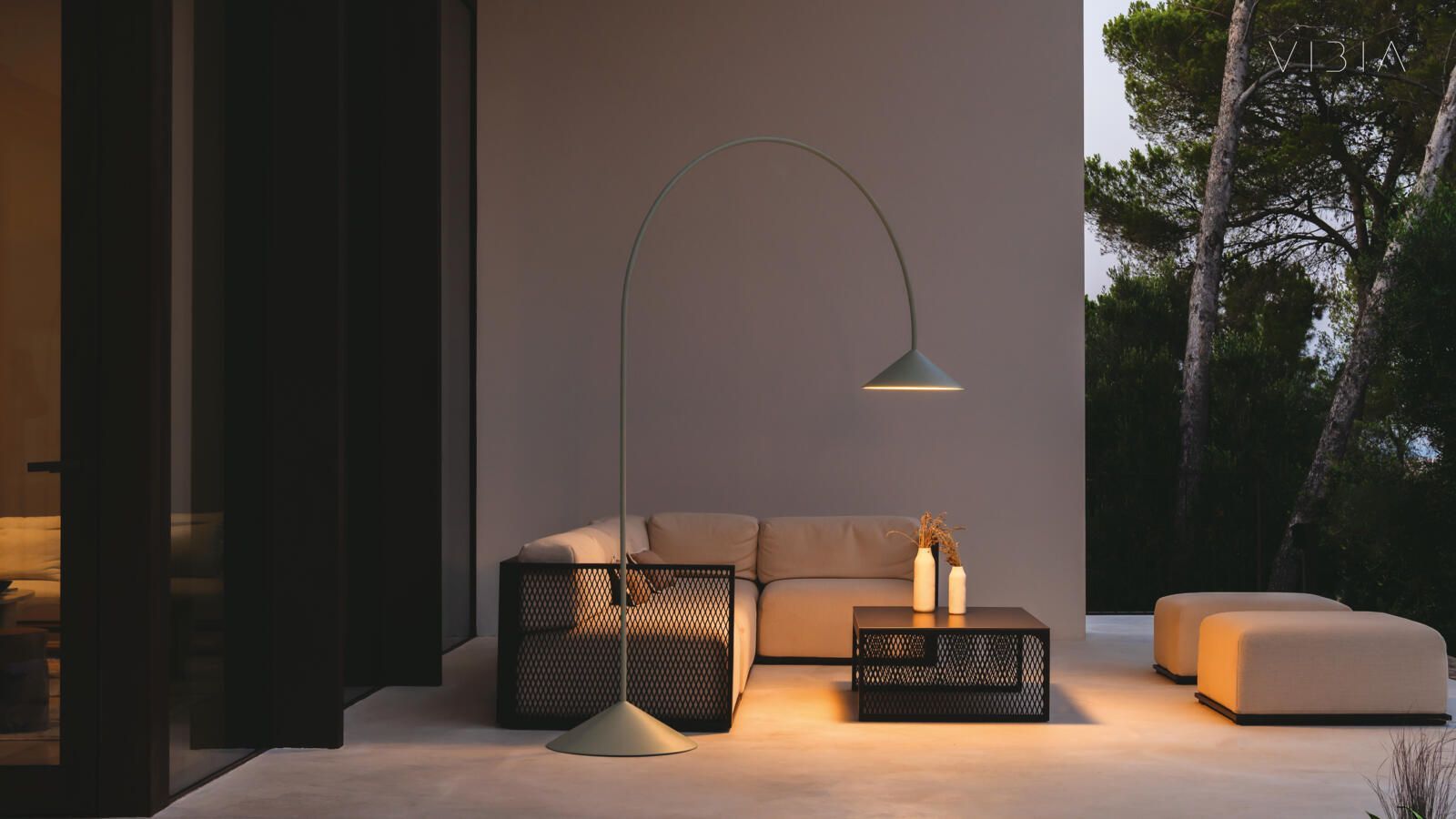 Una escena de un patio al aire libre al atardecer, con una moderna lámpara de pie de arco color beige, un sofá esquinero, una mesa baja y dos otomanas.