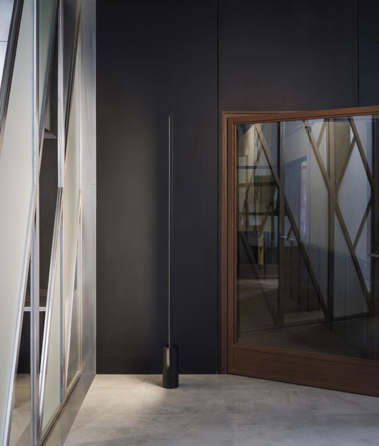 Una lámpara de pie minimalista se alza en una habitación moderna con una pared oscura, una puerta con paneles de cristal y decoración geométrica en la pared.
