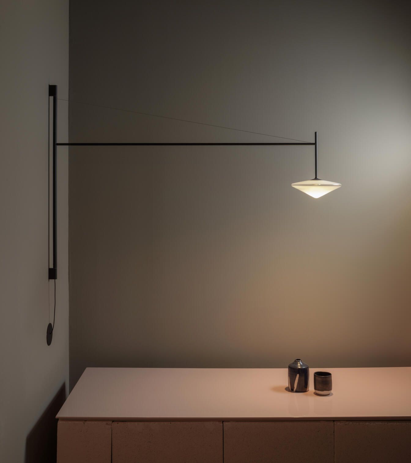 Una lámpara de pared minimalista de color negro con brazo articulado y una pantalla cónica baja y luminosa cuelga sobre una superficie de color rosa pálido.