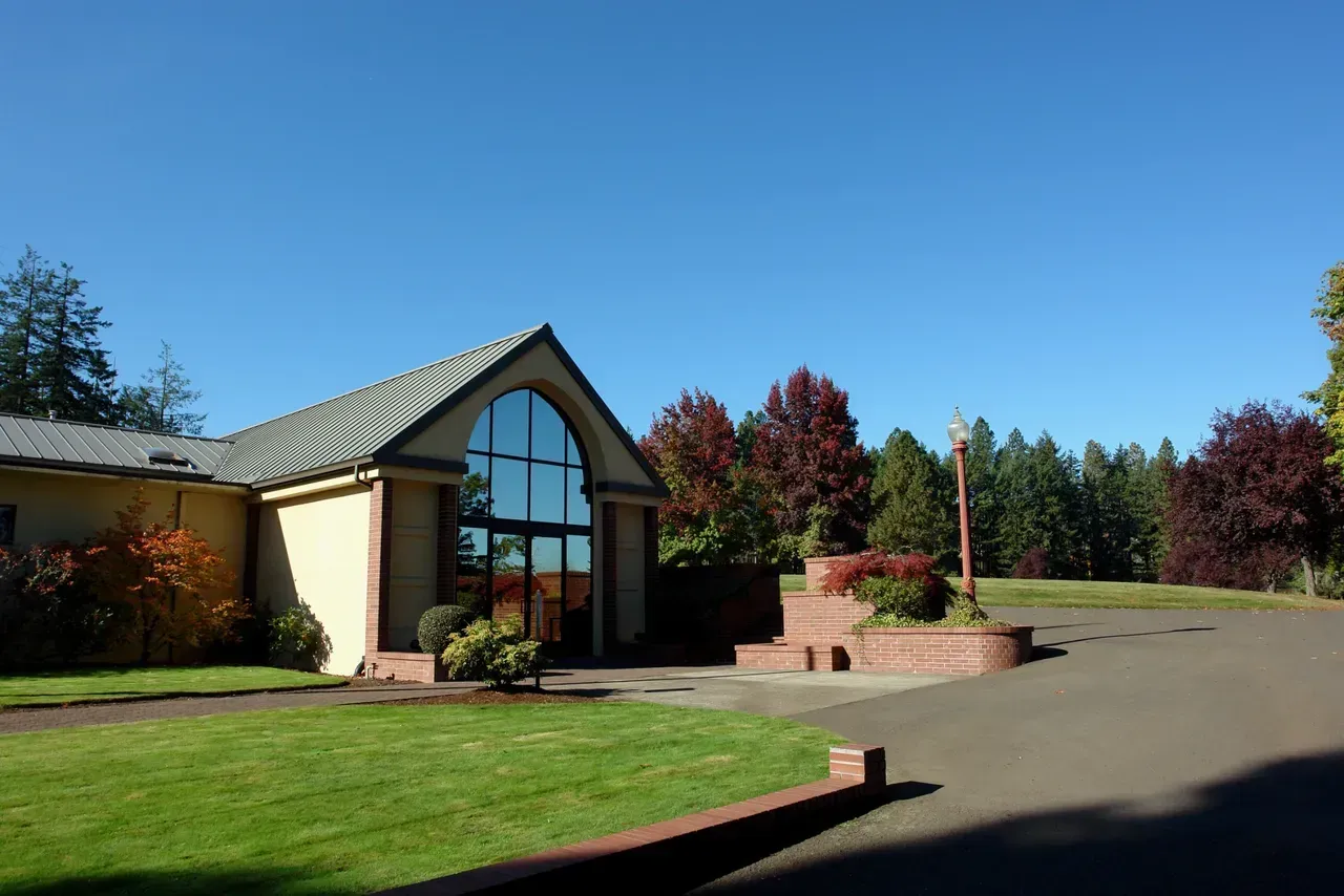 Tan building with large arched windows, green lawn, red-leafed trees, and clear blue sky.