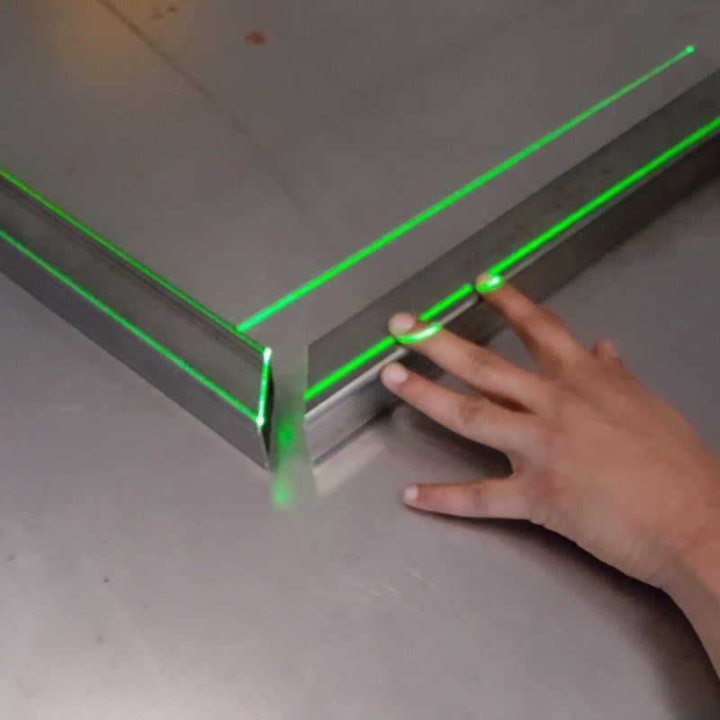 A hand touching a metal corner with green laser lines showing a perfect 90-degree angle.