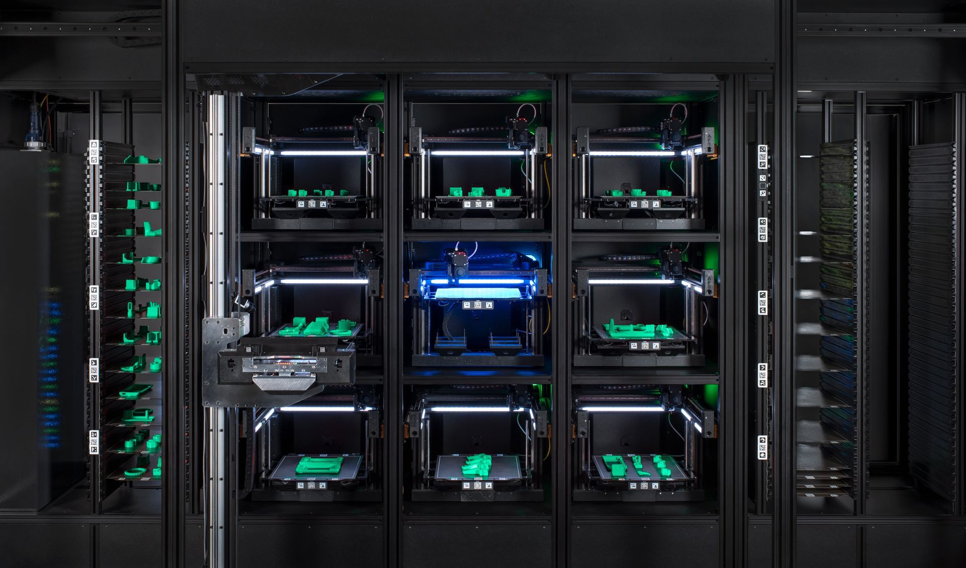 Nine 3D printers in a dark rack, printing green objects. One printer is blue.