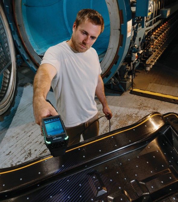Man inspecting carbon fiber part with a handheld device near industrial equipment.