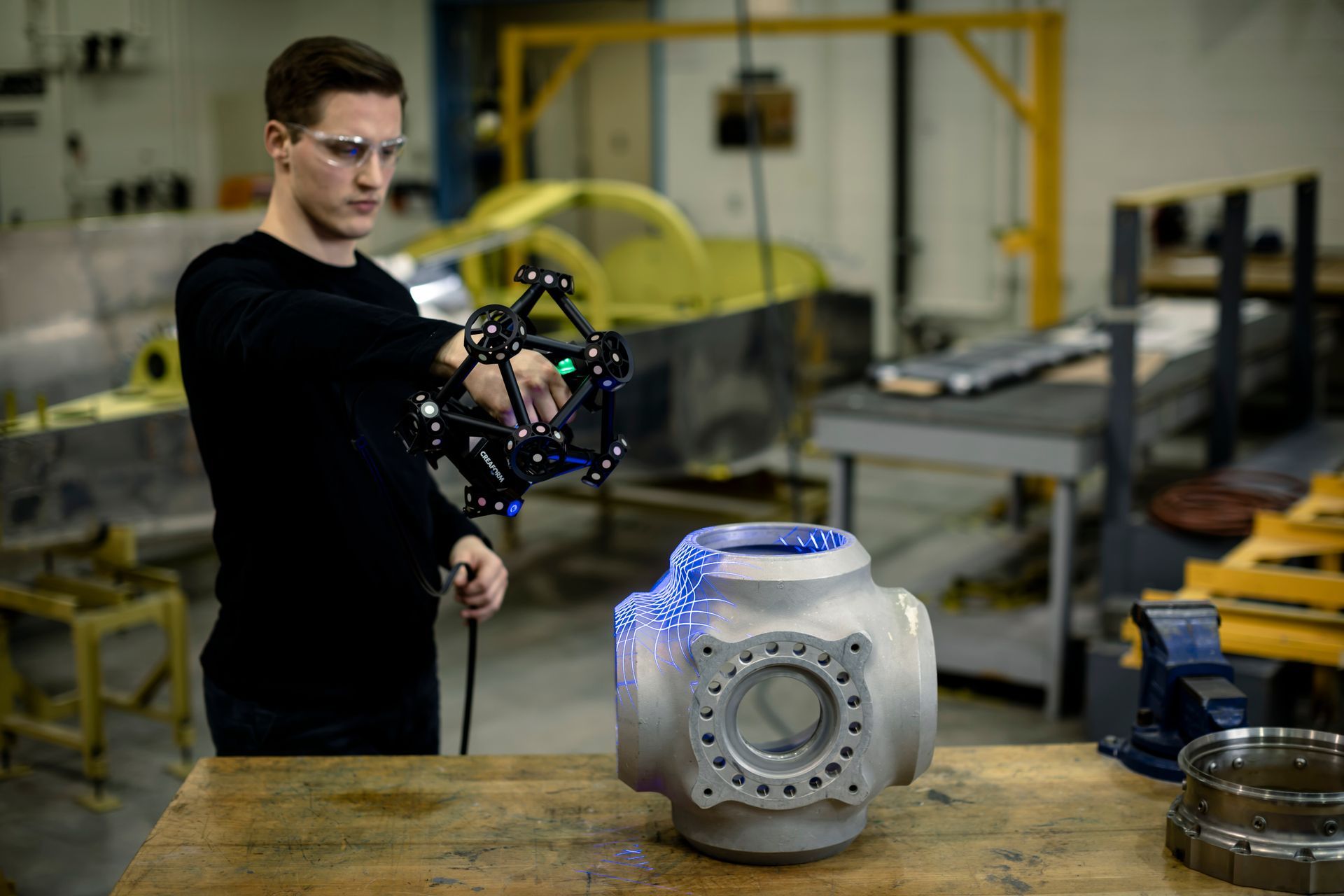 A man is using a 3d scanner to scan a metal object in a factory.