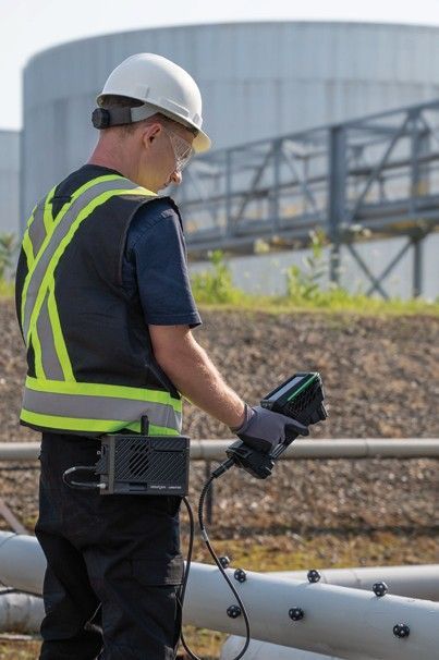 Worker in safety vest using a handheld device to inspect pipes outside of industrial tanks.
