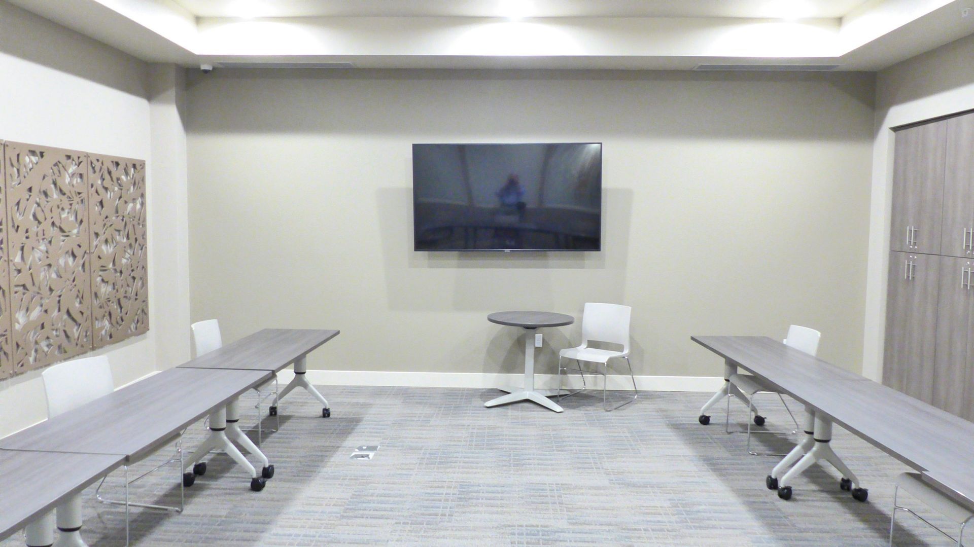 Modern conference room with long tables, white chairs, wall-mounted TV, and patterned carpet